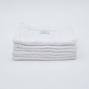 May include: A stack of eight white waffle weave kitchen towels. The top towel has a small white tag with the text "RELIEF GOODS" and "Everyday Essentials" printed on it.
