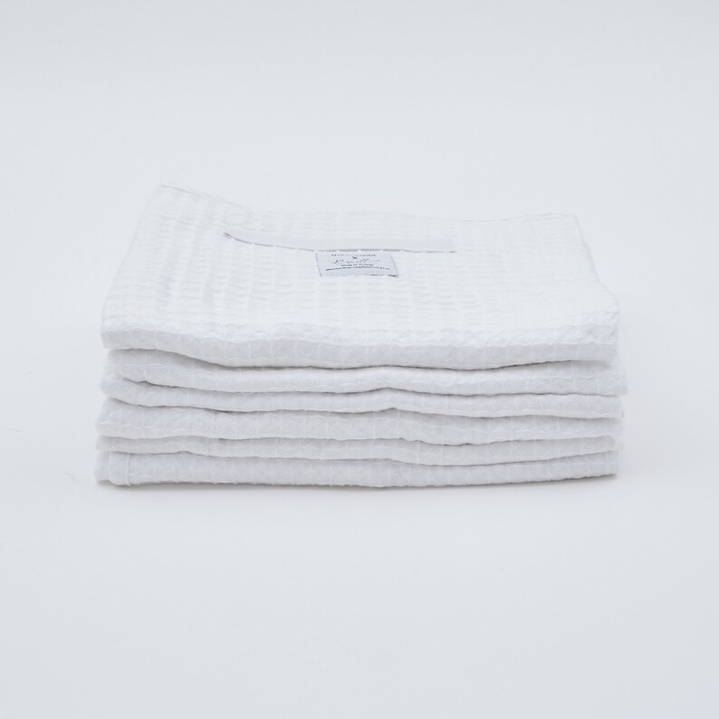 May include: A stack of eight white waffle weave kitchen towels. The top towel has a small white tag with the text "RELIEF GOODS" and "Everyday Essentials" printed on it.