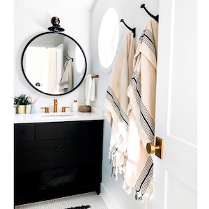 May include: A modern bathroom with a black vanity, gold fixtures, and a round black mirror. Two beige towels with black stripes hang on the wall. A small plant and decorative items sit on the countertop. The room is painted white.