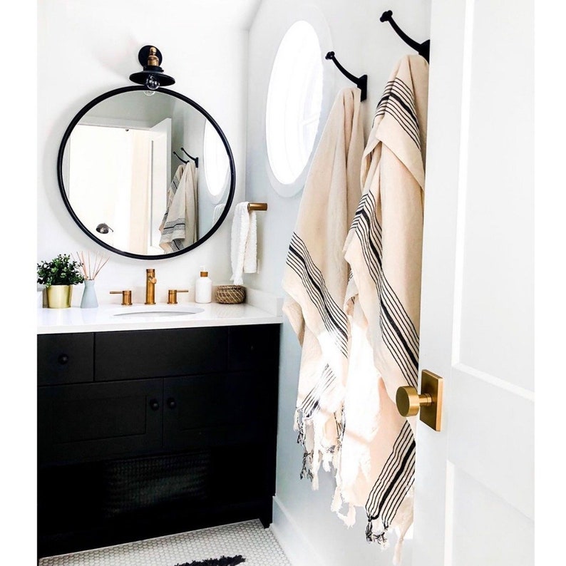 May include: A modern bathroom with a black vanity, gold fixtures, and a round black mirror. Two beige towels with black stripes hang on the wall. A small plant and decorative items sit on the countertop. The room is painted white.