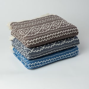 May include: Three woven cotton blankets stacked on top of each other. The top blanket is brown with a white geometric pattern. The middle blanket is gray with a white geometric pattern. The bottom blanket is blue with a white geometric pattern.
