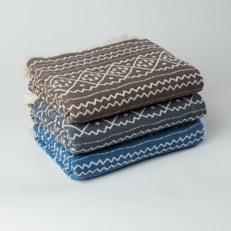 May include: Three woven cotton blankets stacked on top of each other. The top blanket is brown with a white geometric pattern. The middle blanket is gray with a white geometric pattern. The bottom blanket is blue with a white geometric pattern.