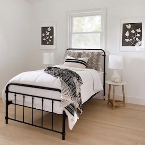 May include: A black metal bed frame with a white duvet and black and white patterned throw blanket. The bed is in a white room with two black and white framed prints on the walls.