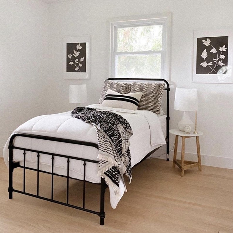 May include: A black metal bed frame with a white duvet and black and white patterned throw blanket. The bed is in a white room with two black and white framed prints on the walls.