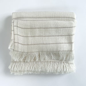 May include: A white striped throw blanket with a fringe border. The blanket is folded in half and has a subtle texture.