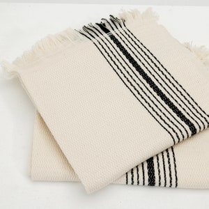 May include: Two off-white square cotton napkins with black stripes and fringe. The napkins have a textured weave and are stacked on top of each other. The black stripes run vertically across the napkins.
