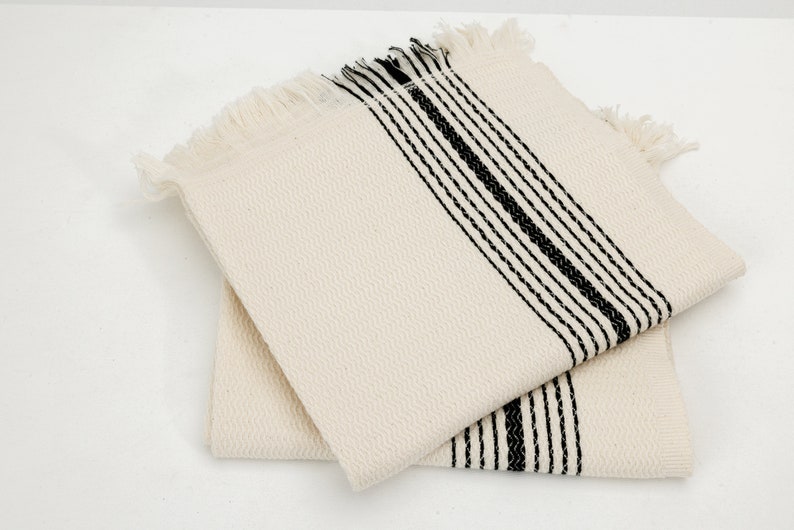 May include: Two off-white square cotton napkins with black stripes and fringe. The napkins have a textured weave and are stacked on top of each other. The black stripes run vertically across the napkins.