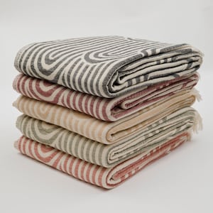 May include: A stack of four patterned throw blankets in various colors, including gray, red, beige, and green. The blankets are folded and stacked on top of each other, with the gray blanket on top.