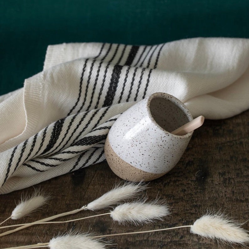 May include: A speckled white and tan ceramic mug with a wooden spoon inside, resting on a wooden surface. A cream-colored towel with black stripes is behind the mug. Dried white flowers are in the foreground.