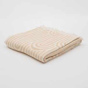 May include: A folded beige throw blanket with a white geometric pattern of stripes and arches. The blanket has a fringe border.