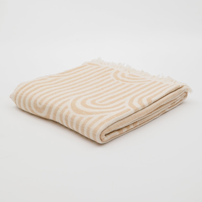 May include: A folded beige throw blanket with a white geometric pattern of stripes and arches. The blanket has a fringe border.