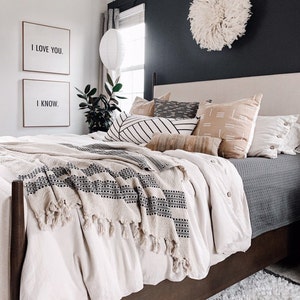 May include: A bedroom scene featuring a bed with a beige headboard and a variety of decorative pillows. The bed is covered with a cream-colored duvet and a patterned throw blanket. Two framed prints on the wall read "I LOVE YOU." and "I KNOW."