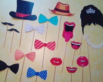 May include: A collection of photo booth props on sticks, including a black top hat, a tan fedora, a black hairstyle with a tiara, a white pearl necklace, various bow ties in solid and patterned colours, two black moustaches, and four sets of red lips.