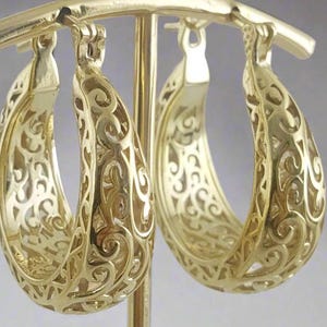 May include: A pair of gold-colored hoop earrings with an intricate, openwork filigree design. The earrings are teardrop-shaped and hang from a gold-colored stand. The filigree pattern features swirling, floral-like details.