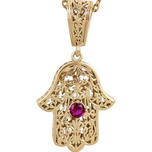 May include: A gold-colored Hamsa hand pendant necklace. The pendant features intricate openwork and a central, round, red gemstone. The necklace chain is also gold-colored, and the pendant hangs from a decorative bail.