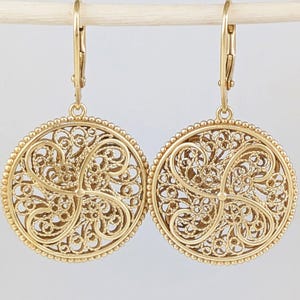 May include: A pair of gold-colored filigree earrings. Each earring features a circular design with intricate, swirling patterns and a beaded border. The earrings hang from lever-back closures.