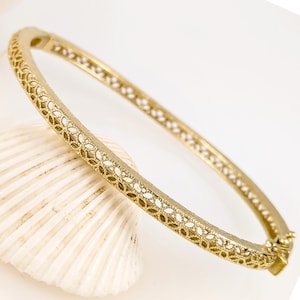 May include: A gold-colored bangle bracelet with an intricate openwork design. The bracelet is hinged and has a secure clasp. The bracelet is resting on a white seashell against a white background. Jewelry.