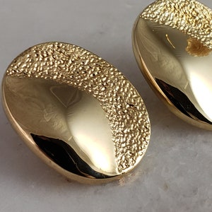 May include: Two gold-coloured oval earrings. Each earring features a smooth, polished surface on one side and a textured, granular design on the other. The earrings are presented on a white marble surface.