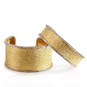 Puede incluir: Dos brazaletes anchos tipo puño con un acabado dorado texturizado en la superficie exterior. Cada brazalete está bordeado por finos bordes plateados incrustados con pequeñas piedras claras que captan la luz, creando un contraste distintivo.