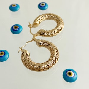 May include: Gold filigree hoop earrings with a delicate, intricate design. The earrings are displayed on a white surface with several blue evil eye charms.