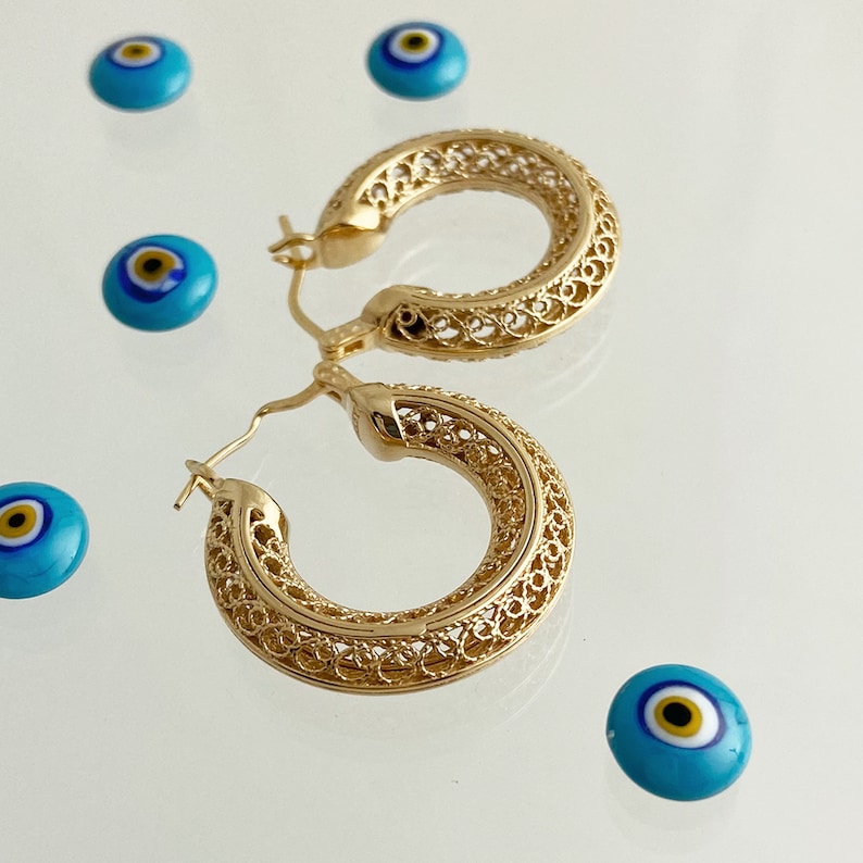 May include: Gold filigree hoop earrings with a delicate, intricate design. The earrings are displayed on a white surface with several blue evil eye charms.