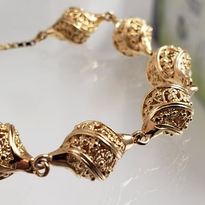 Puede incluir: Un brazalete de oro con un diseño delicado e intrincado. El brazalete está formado por una serie de eslabones conectados y adornados que se asemejan a pequeñas conchas retorcidas.