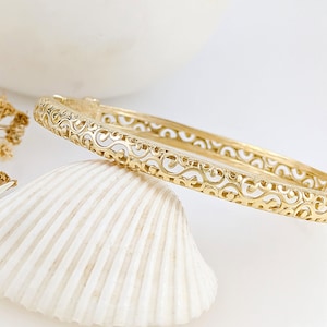 May include: A gold-coloured bangle bracelet with an intricate openwork design. The bracelet is resting on a white seashell, with a blurred white background. The bracelet's pattern features swirling, symmetrical shapes.