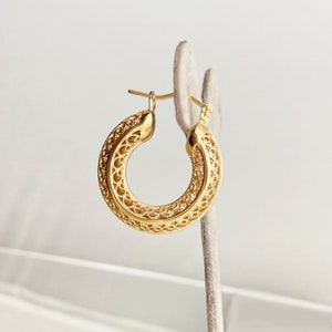 May include: Gold hoop earrings with a delicate, intricate filigree design. The earrings are displayed on a white stand.