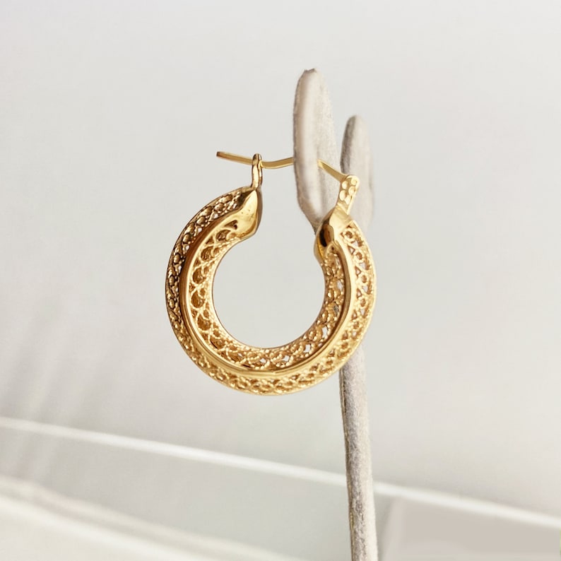 May include: Gold hoop earrings with a delicate, intricate filigree design. The earrings are displayed on a white stand.