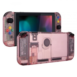 Puede incluir: Una consola Nintendo Switch transparente rosa con botones coloridos en el joy-con izquierdo. La consola se muestra tanto en modo portátil como en modo de sobremesa.