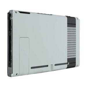 Peut inclure: Une console Nintendo Switch blanche avec un design NES noir et gris. La console a une bordure noire autour des bords et un logo NES gris au dos.