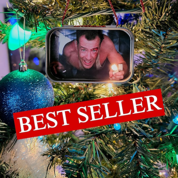 May include: A rectangular metal ornament featuring a printed image of a man with a bloody face crawling through a vent while holding a lit lighter. The ornament hangs from a red string and includes the text BEST SELLER in a red banner.