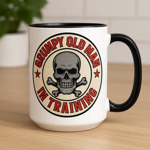 May include: White ceramic mug with a black handle and rim. The mug features a circular design with a gray skull and crossbones, surrounded by the text "GRUMPY OLD MAN IN TRAINING" in red.