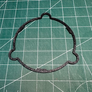 May include: A black, textured, circular gasket with a unique shape, featuring a curved top and two side protrusions. The gasket is positioned on a green cutting mat with white grid lines, providing a contrasting background.