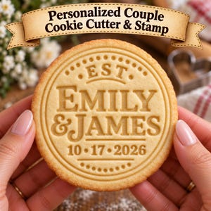 May include: A round, light-tan cookie featuring an embossed design with the text "EST. EMILY & JAMES" and the date "10-17-2026" centered within a decorative circular border. A banner above the cookie displays the text "Personalized Couple Cookie Cutter & Stamp".