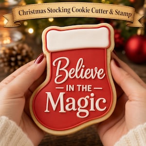May include: A Christmas stocking-shaped cookie with red icing and a white cuff. The text "Believe IN THE Magic" is written in white icing across the center. A banner at the top reads "Christmas Stocking Cookie Cutter & Stamp" in dark text.