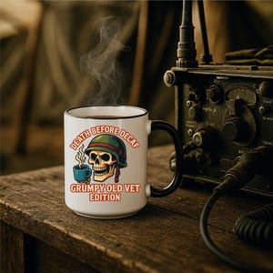 May include: A white coffee mug with a black handle and rim. The mug features a skull wearing a green helmet and holding a blue cup, with the text "DEATH BEFORE DECAF" and "GRUMPY OLD VET EDITION". Steam rises from the coffee.