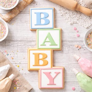 May include: Square-shaped cookies spelling out "BABY" with pastel-colored icing. The letters are blue, green, orange, and pink, each on a white background. The cookies are arranged on a light wooden surface, with sprinkles and pastry tools nearby.