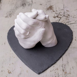 May include: A white plaster cast of two hands holding each other, with a silver ring on one of the fingers. The cast is displayed on a black heart-shaped slate.