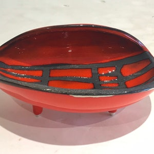 May include: A red ceramic bowl with a black geometric design and three small legs. The bowl is oval shaped and has a glossy finish.