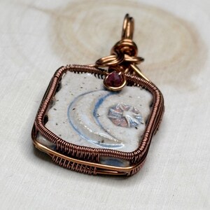 May include: Handmade pendant with a square ceramic tile, displaying a crescent moon and star design in blue and white. The tile is encased in copper wire, with a small red gemstone.
