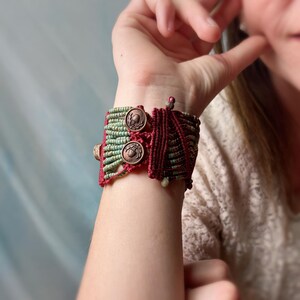 May include: A handmade macrame bracelet in shades of green and burgundy. The bracelet features two copper-colored buttons with a detailed design. The intricate knotting and color combination create a unique accessory.