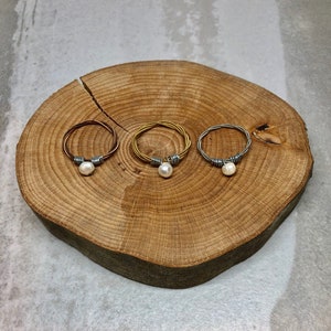 Puede incluir: Tres anillos de plata con una perla unida a cada anillo. Los anillos están hechos de cuerdas de guitarra y se muestran sobre una superficie de madera.