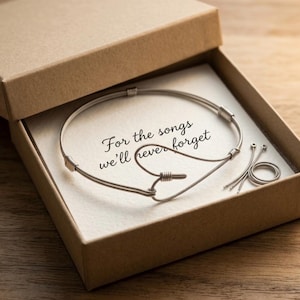 Könnte beinhalten: Ein silberfarbenes Armband mit herzförmigem Design, präsentiert in einer braunen Geschenkbox. Das Armband ist aus Draht gefertigt und trägt die Aufschrift "For the songs we'll never forget" auf der Karte in der Box. Zusätzliche Drahtstücke sind enthalten.