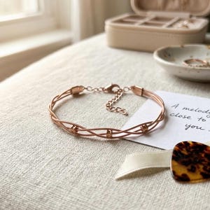 May include: Rose gold bracelet with a woven design. The bracelet is on a neutral-colored surface, with a small card that reads "A melody close to you." A jewelry box and dish with rings are in the background.