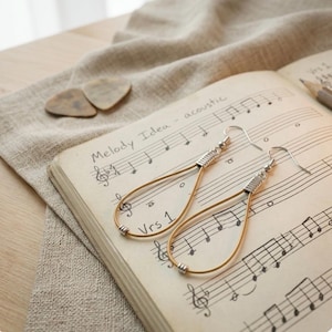 Könnte beinhalten: Ohrringe in Tropfenform mit goldfarbenem Drahtrahmen und silbernen Akzenten. Sie liegen auf einem alten Notenblatt mit dem Text "Melody Idea - acoustic". Zwei Plektren und ein Bleistift sind ebenfalls zu sehen.