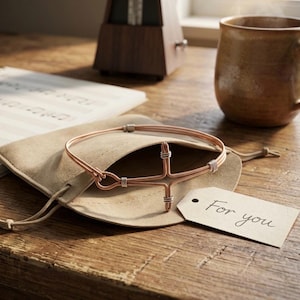 Puede incluir: Una pulsera de alambre color cobre con diseño de cruz, sobre una bolsa beige. Una etiqueta con "For you" está adjunta. La pulsera está sobre una superficie de madera, con una taza y partituras en el fondo.