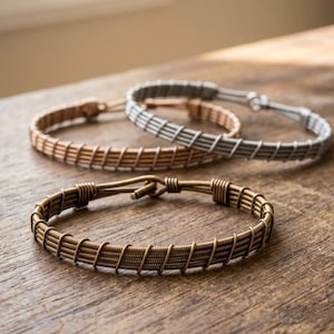 Könnte beinhalten: Drei handgefertigte Drahtarmbänder in Kupfer-, Silber- und Bronzetönen. Die Armbänder werden auf einer Holzoberfläche präsentiert, die ihre komplizierten Designs und metallischen Oberflächen hervorhebt. Dies sind Schmuckstücke.