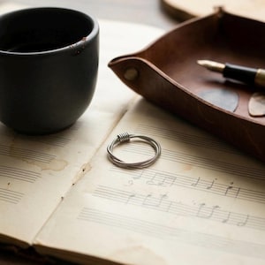 May include: A silver ring crafted from intertwined metal wires sits on aged sheet music. A black ceramic cup and a brown leather tray with a pen and guitar plectrums are also visible, creating a musician's aesthetic.