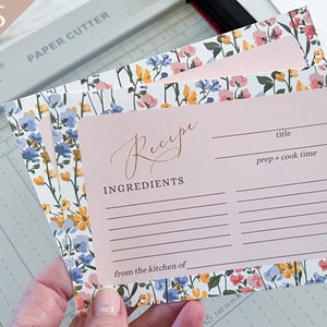 Puede incluir: Un conjunto de tarjetas de recetas imprimibles con un diseño floral. Las tarjetas son de color rosa con líneas blancas para escribir. El texto "Receta" está escrito en cursiva en la tarjeta superior. El texto "Ingredientes" está escrito debajo de la palabra "Receta". El texto "de la cocina de" está escrito en la parte inferior de la tarjeta. El texto "título" está escrito en la esquina superior derecha de la tarjeta. El texto "tiempo de preparación + tiempo de cocción" está escrito debajo de la palabra "título". Las tarjetas están sobre una superficie blanca con una cortadora de papel.